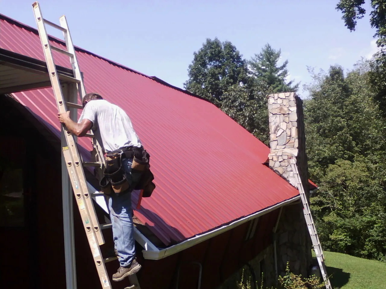 Roofer on a ladder beside a red metal roof and stone chimney — Soffit & Fascia Repair by Roofing Brothers in Mount Olivet